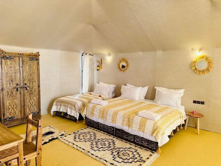 Sunset Desert Camps cozy tent interior with two beds featuring yellow and white patterned blankets, wooden wardrobe, and rustic décor