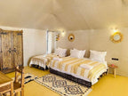 Sunset Desert Camps cozy tent interior with two beds featuring yellow and white patterned blankets, wooden wardrobe, and rustic décor
