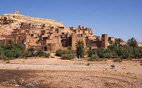 Ancient mud-brick Kasbah with terracotta walls and palm trees in a desert landscape, illustrating Taxi rent from Merzouga to Fes service.