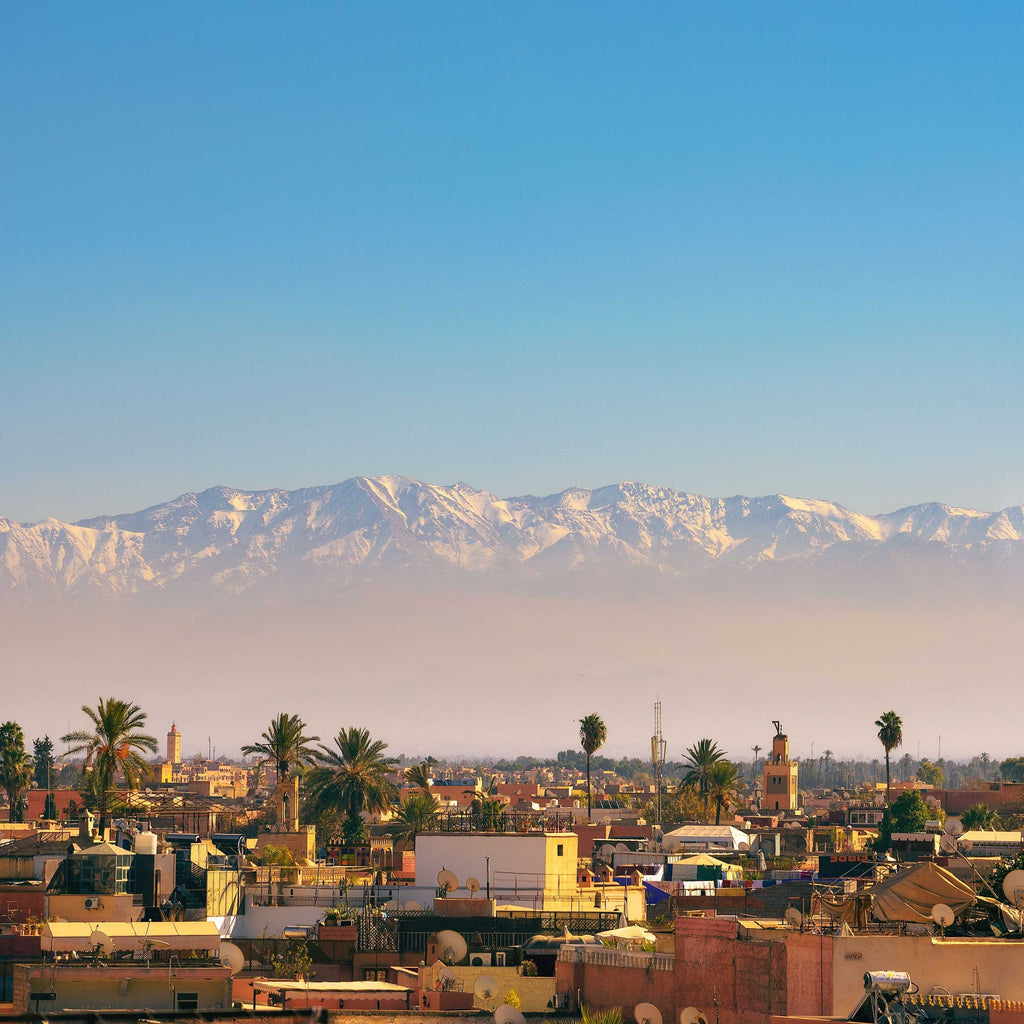 Cityscape of Marrakech with warm-toned buildings, palm trees, and snowy Atlas Mountains in the background, perfect for Taxi rent from Merzouga to Fes travel.