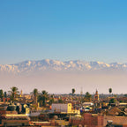 Cityscape of Marrakech with warm-toned buildings, palm trees, and snowy Atlas Mountains in the background, perfect for Taxi rent from Merzouga to Fes travel.