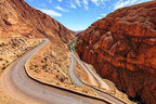 Scenic winding asphalt road through rugged reddish-brown rocky mountains in a dry desert landscape, ideal for Taxi rent from Merzouga to Fes.