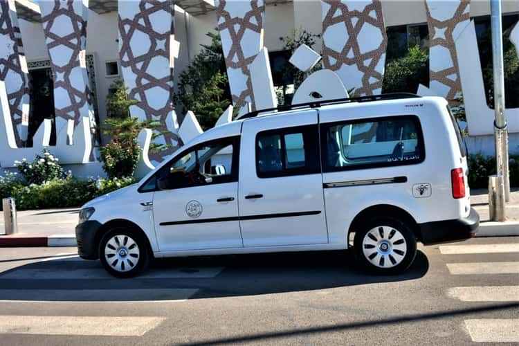 White Volkswagen van taxi with black accents and tinted windows parked on a street near patterned building, ideal for Taxi rent from Merzouga to Fes.