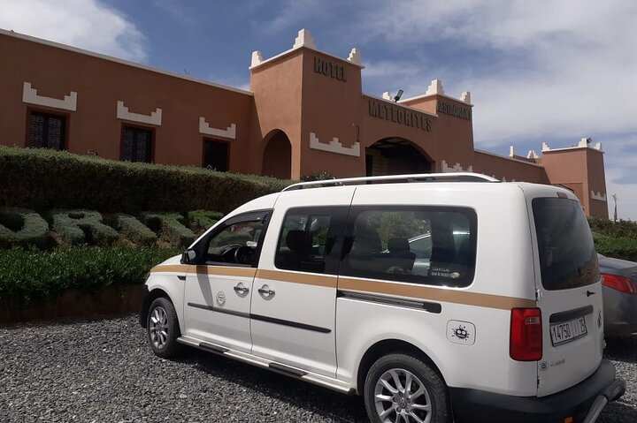 White taxi van with black and gold stripes, roof rack, and tinted windows parked in front of a terracotta hotel building, ideal for Taxi rent from Merzouga to Fes.