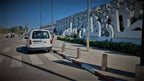 White taxi van driving on a paved road with red and white curb markings, modern decorative white wall and green plants alongside, Taxi rent from Merzouga to Fes.
