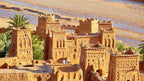 Traditional mud-brick Kasbah buildings with intricate patterns and palm trees under sunlight near a river valley, illustrating Taxi Rental from Merzouga to Ouarzazate scenery.