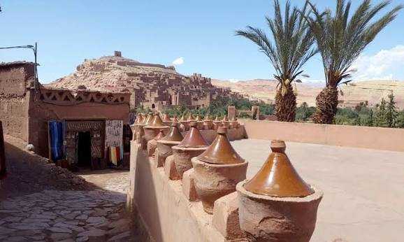 Traditional tan clay pottery jars with conical lids on a mud-walled terrace near palm trees and ancient desert village ruins, illustrating Taxi Rental from Merzouga to Ouarzazate.