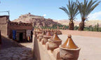 Traditional tan clay pottery jars with conical lids on a mud-walled terrace near palm trees and ancient desert village ruins, illustrating Taxi Rental from Merzouga to Ouarzazate.