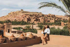 Scenic desert landscape with traditional mud-brick buildings and a woman sitting on a tan wall under a palm tree, promoting Taxi Rental from Merzouga to Ouarzazate.