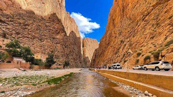 Scenic narrow canyon road with light-colored stone cliffs, a clear stream, and white cars parked on the roadside, illustrating Taxi Rental from Merzouga to Ouarzazate travel route.