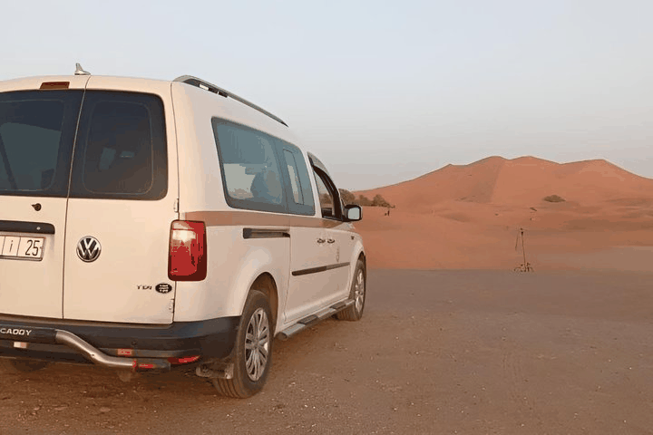 White Volkswagen SUV parked on sandy desert terrain near large orange sand dunes, ideal for Taxi Rental from Merzouga to Ouarzazate.