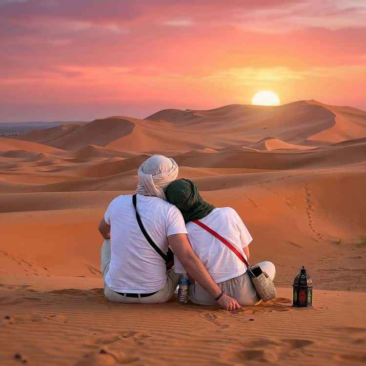 Couple sitting on golden sand dunes at sunset with traditional headscarves, highlighting a serene moment for Taxi Transfer from Errachidia Airport.