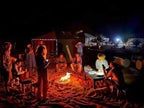 Group of people gathered around an open campfire at night on sand near desert tents, illuminated by warm light, Taxi Transfer from Errachidia Airport trip scene.