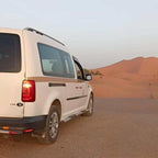 White van taxi with tinted rear windows parked on a desert road near sand dunes during sunset for Taxi Transfer from Errachidia Airport.