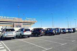 Parking lot with a row of white, silver, and black SUVs parked outside Moulay Ali Cherif Airport under clear blue sky for Taxi Transfer from Errachidia Airport.