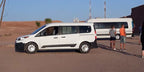 White and black passenger van parked on desert terrain with people nearby, suitable for Taxi Transfer from Errachidia Airport service