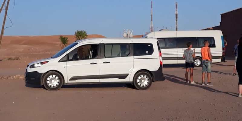 White and black passenger van parked on desert terrain with people nearby, suitable for Taxi Transfer from Errachidia Airport service