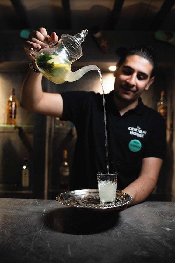 Bartender pouring traditional Moroccan mint tea from a glass teapot into a small glass on a tray at The Central House Marrakech Medina