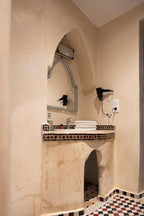 Bathroom vanity with beige plaster walls, decorative mosaic tile trim, arched niche, mirror, hairdryer, and folded white towels in The Central House Marrakech Medina