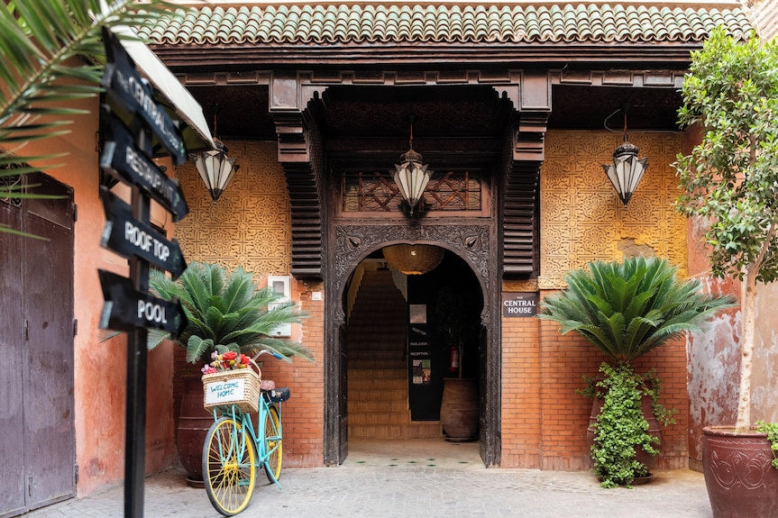 The Central House Marrakech Medina entrance with intricate wooden archway, terracotta walls, potted plants, and a bicycle with a flower basket