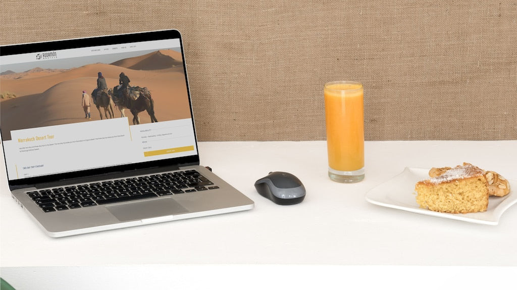 Laptop displaying Moroccan desert tour website, wireless mouse, glass of orange juice, and plate with cake and pastry on white table