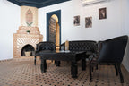 The Central House Marrakech Medina living room with black leather tufted sofas, ornate wooden table, patterned tiled floor, and decorative brick fireplace