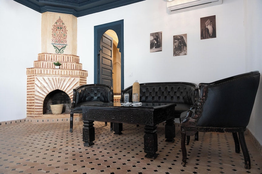 The Central House Marrakech Medina living room with black leather tufted sofas, ornate wooden table, patterned tiled floor, and decorative brick fireplace