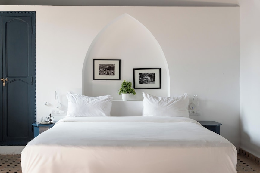 The Central House Marrakech Medina bedroom with a large white bed, white arched wall niche, two black and white framed photos, and blue door