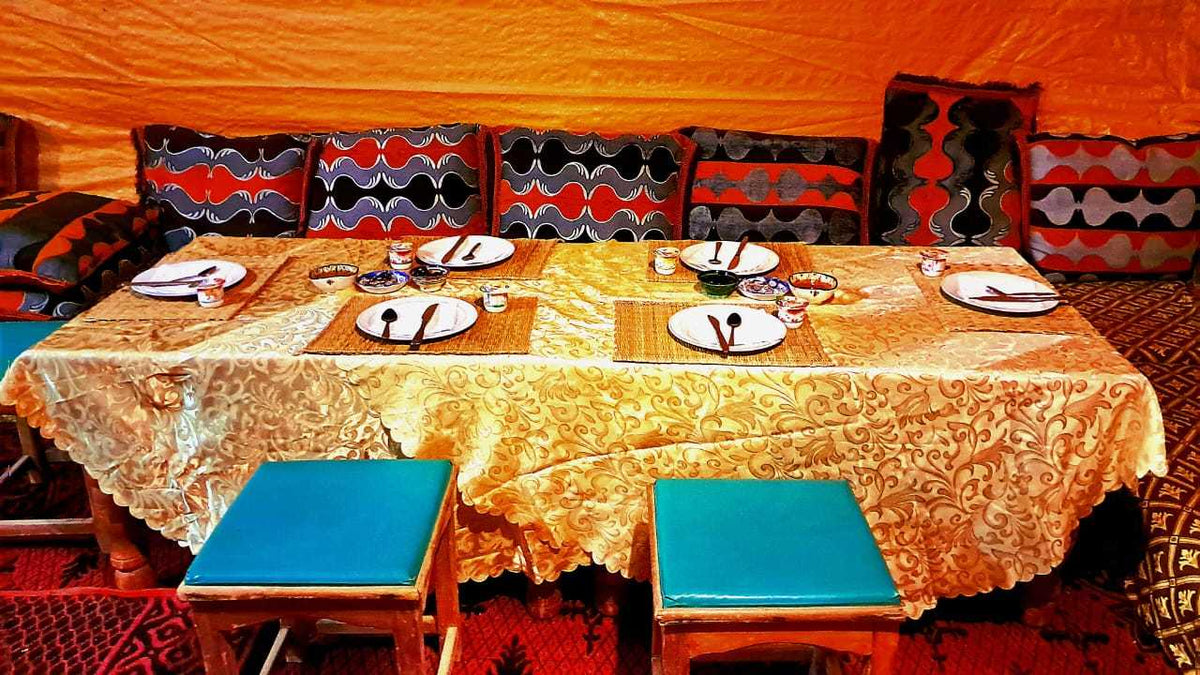 Tigmi Standard Desert Camp interior with ornate golden tablecloth, colorful patterned cushions, and blue cushioned stools