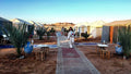 Tents with white canvas and blue accents set in a desert with rugs, small wooden stools, plants, and people at Tigmi Standard Desert Camp.