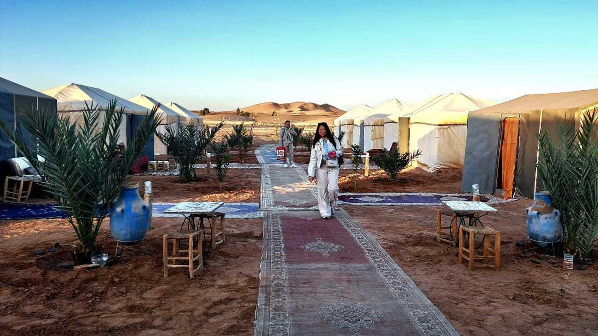 Tents with white canvas and blue accents set in a desert with rugs, small wooden stools, plants, and people at Tigmi Standard Desert Camp.