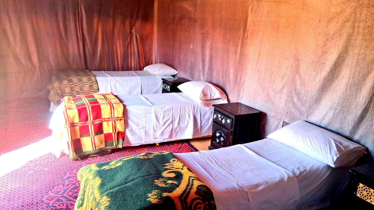 Tigmi Standard Desert Camp interior with three single beds, white sheets, colorful patterned blankets, and dark wood bedside tables in a fabric tent