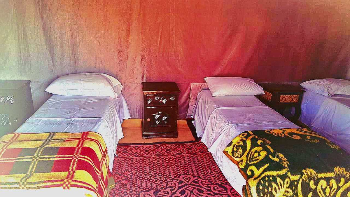 Tigmi Standard Desert Camp interior showing three single beds with white linens and patterned blankets, traditional rugs, and wooden bedside tables
