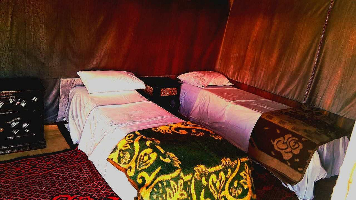 Tigmi Standard Desert Camp interior with two single beds dressed in white linens and patterned blankets inside a brown tent