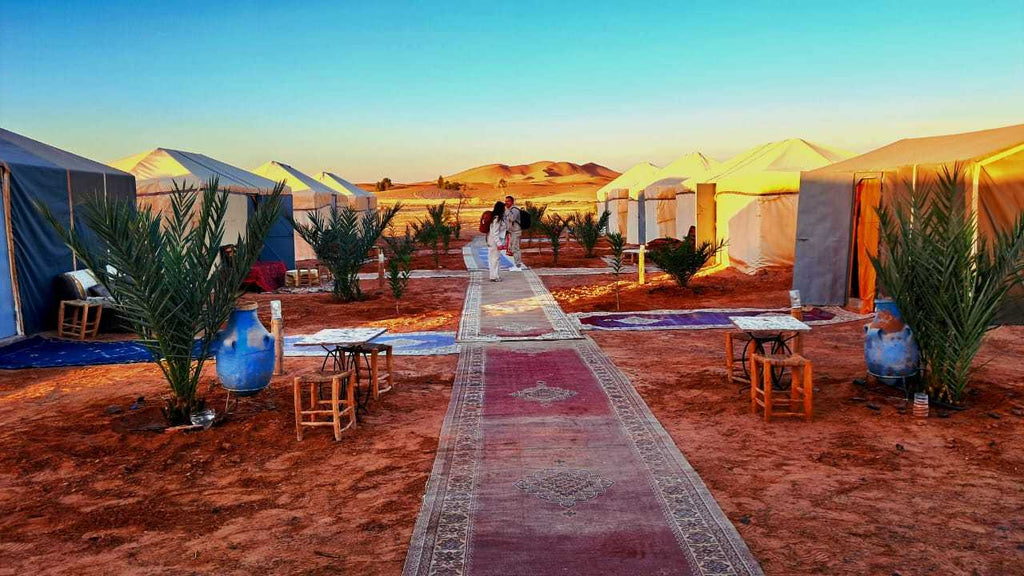 Tigmi Standard Desert Camp with white canvas tents, red sandy ground, decorative rugs, small wooden stools, and desert plants under a clear sky