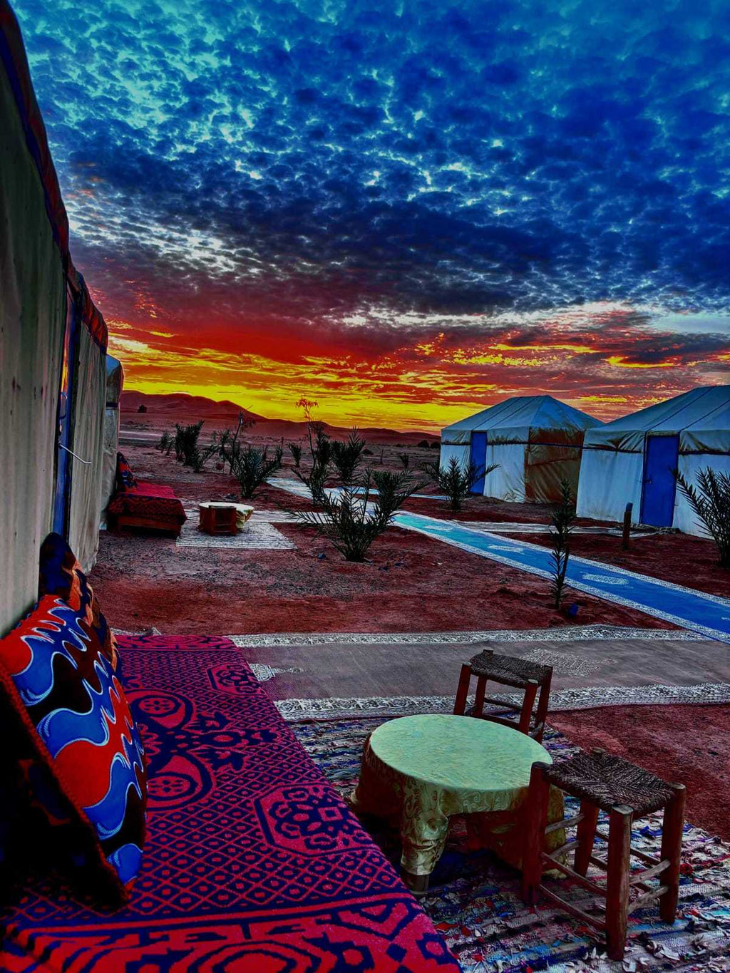 Tigmi Standard Desert Camp with traditional white tents, colorful patterned carpets and cushions, wooden stools under a vibrant sunset sky