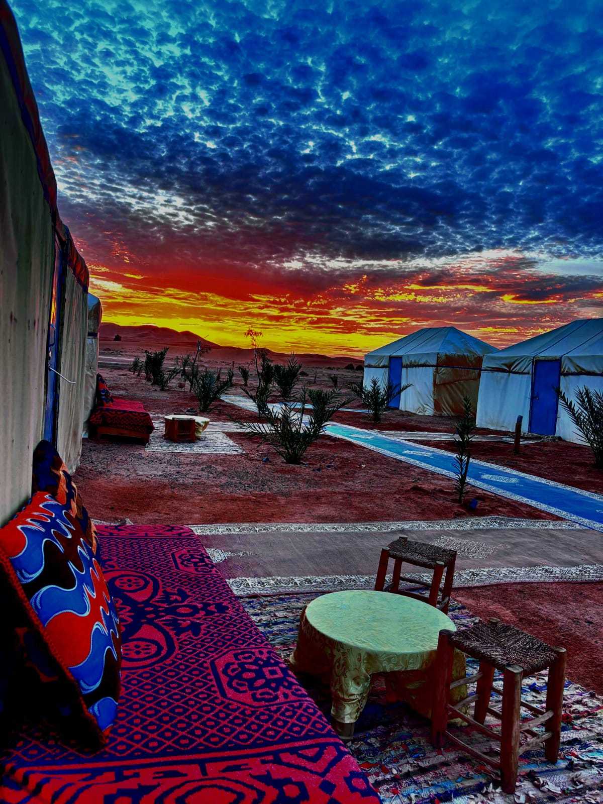 Tigmi Standard Desert Camp with traditional white tents, colorful patterned carpets and cushions, wooden stools under a vibrant sunset sky