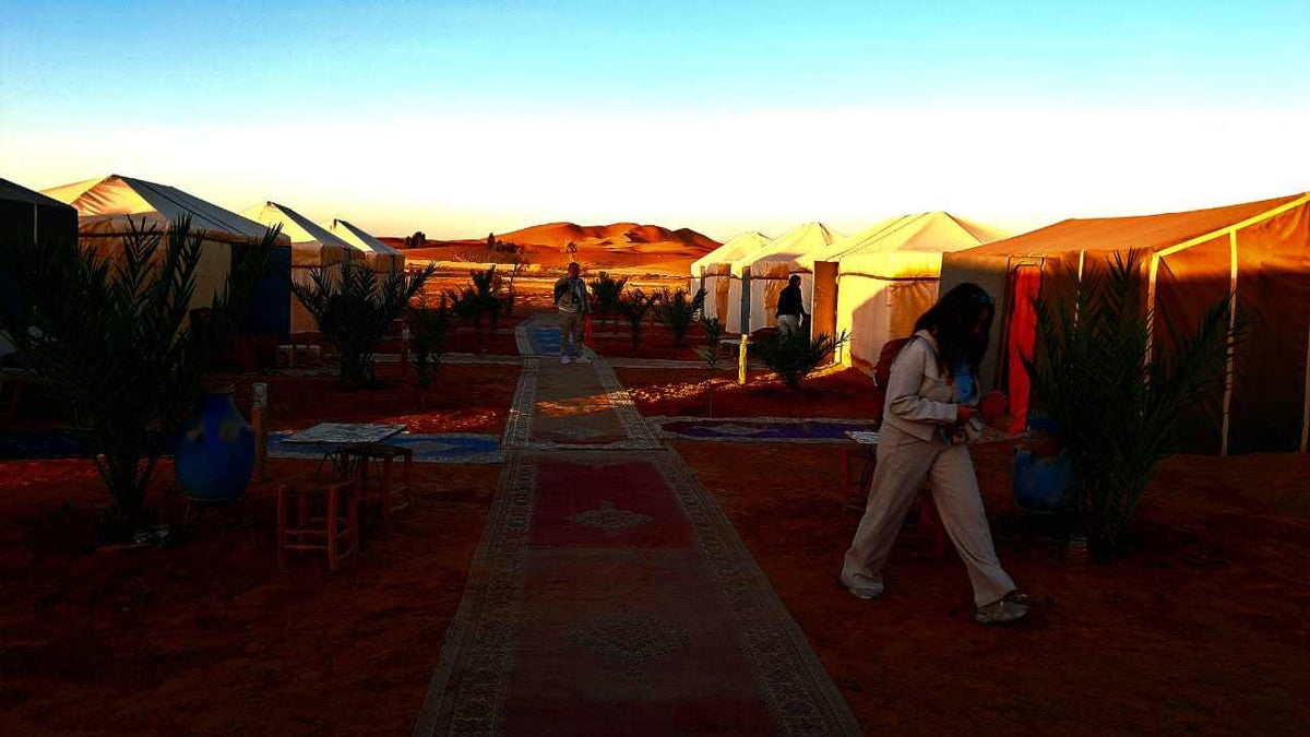 Tigmi Standard Desert Camp with white canvas tents and carpets lining sandy path in Moroccan desert under blue sky