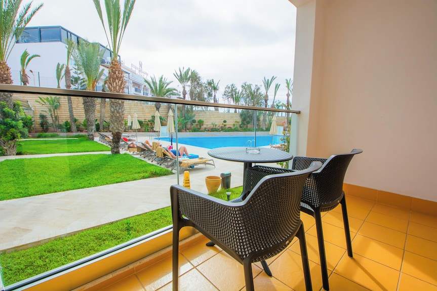 Outdoor balcony at Timoulay Hotel & Spa Agadir overlooking a swimming pool, green lawn, and palm trees with black woven chairs and round table