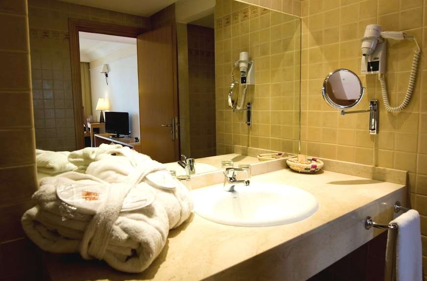 Bathroom countertop with beige tiles, white sink, wall-mounted magnifying mirror, hairdryer, and folded white bathrobes at Timoulay Hotel & Spa Agadir