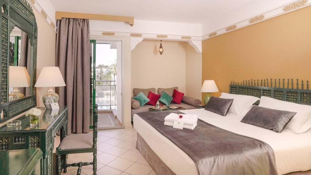 Timoulay Hotel & Spa Agadir bedroom with beige walls, green carved wooden furniture, large bed with grey accents, and seating area with colorful cushions