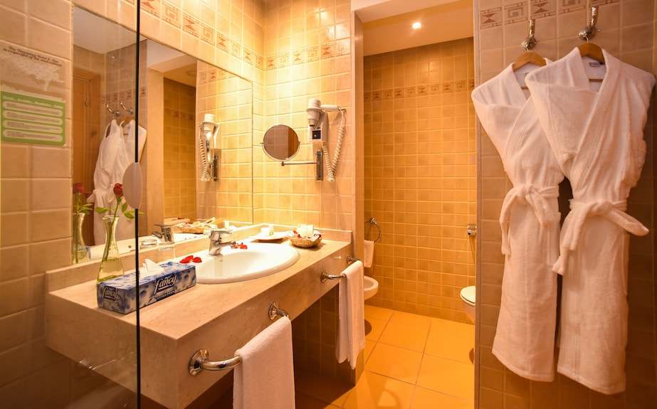 Bathroom with beige tiled walls, cream stone countertop, white towels, two hanging white bathrobes, and wall-mounted hairdryer in Timoulay Hotel & Spa Agadir