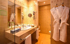 Bathroom with beige tiled walls, cream stone countertop, white towels, two hanging white bathrobes, and wall-mounted hairdryer in Timoulay Hotel & Spa Agadir