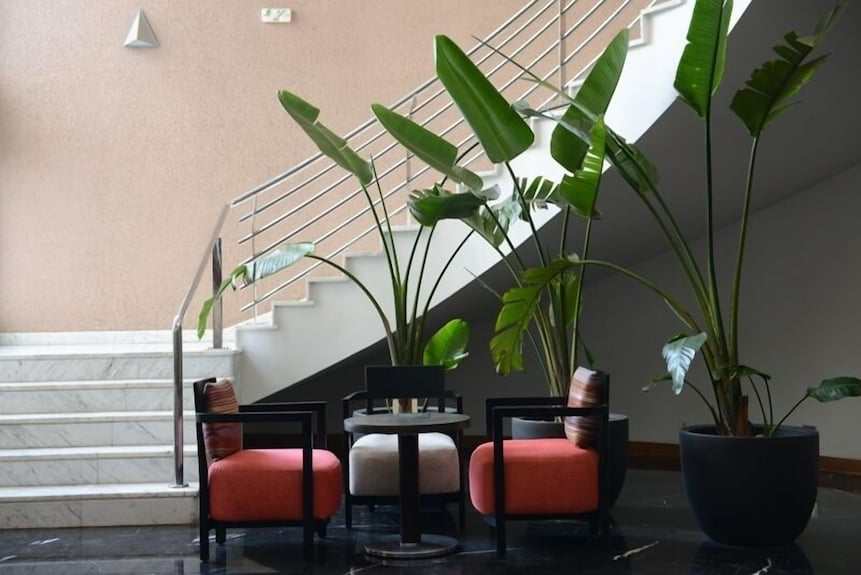 Seating area with two coral upholstered armchairs, a beige ottoman, black round table, and large green potted plants near marble stairs