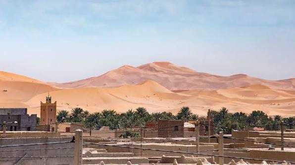 Tinghir to Merzouga view showing golden desert dunes behind a traditional village with palm trees and clay buildings under a clear blue sky