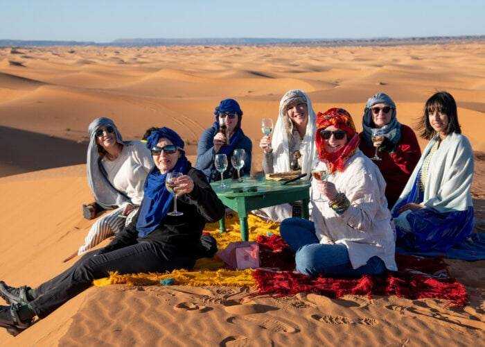 Group of seven people wearing colorful headscarves and blankets enjoying drinks seated around a green table in the golden sand dunes of Merzouga desert