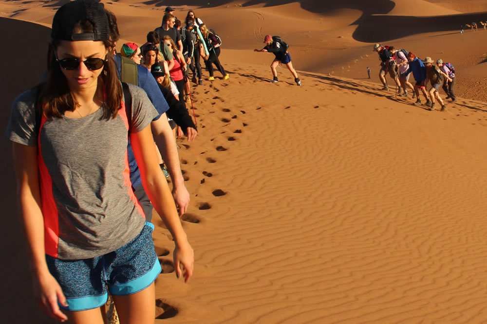 Group of tourists trekking up golden sand dunes under bright sunlight with footprints in the sand, illustrating Tinghir to Merzouga journey