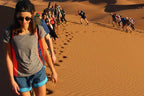 Group of tourists trekking up golden sand dunes under bright sunlight with footprints in the sand, illustrating Tinghir to Merzouga journey