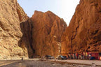 Scenic narrow gorge with towering red rock cliffs, small group of people, parked cars, and colorful market stalls in a desert landscape