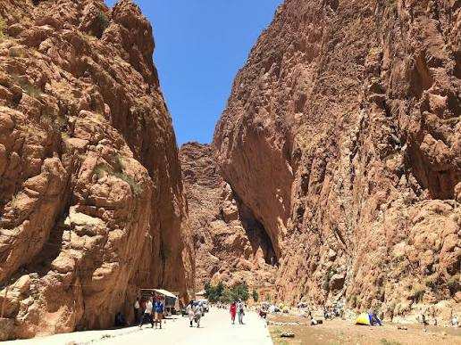 Todra Gorge canyon with steep reddish-brown rocky cliffs and groups of people walking on a paved path, Tinghir to Merzouga trip.